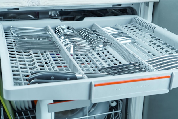 Closeup of grey compartment tray of open dishwasher machine with cutlery set of knives spoons forks tea spoons