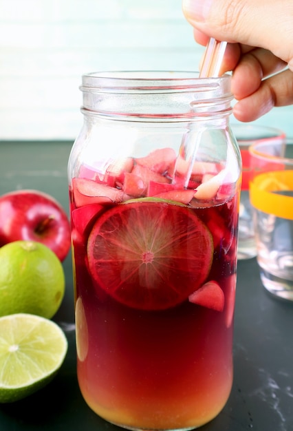 Closeup of a Hand Mixing Red Wine Sangria in Grass Bottle with Blurry Ingredients in Background