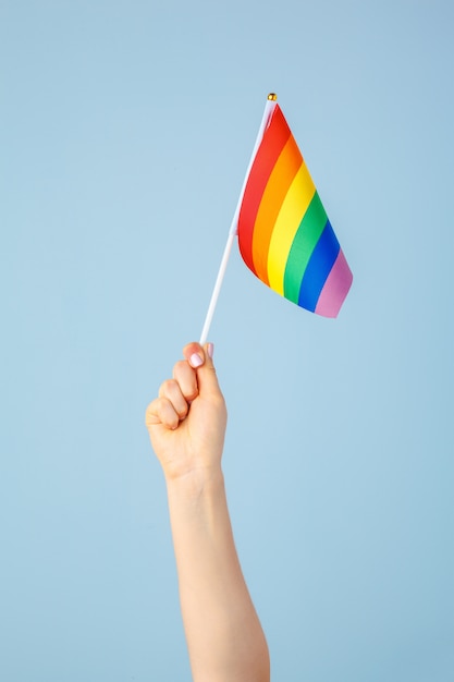 Closeup of a hand waving a small rainbow flag against a light  blue  