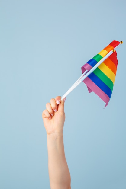 Closeup of a hand waving a small rainbow flag against a light  blue  