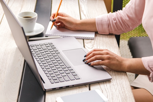 Mani del primo piano della donna di affari in camicia di rosa pastello che lavora al computer portatile e che fa gli avvisi in suo taccuino sulla tavola di legno con una tazza di caffè