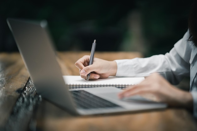 Closeup hands of business woman working on laptop and using pen making notices in her notebook on wooden table with a cup of coffee Business concept