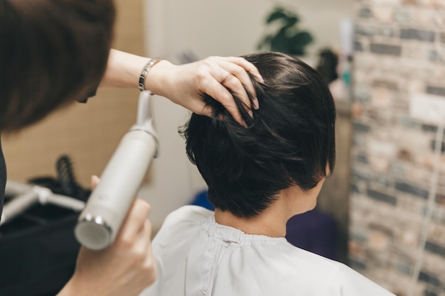 ヘアドライヤーの短いヘアカットとスタイリングで女性の髪を乾燥させる美容師の手の接写