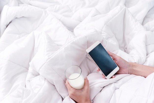 Closeup of hands holding mobile and milk glass on bed in the morning. Technology for life concept.