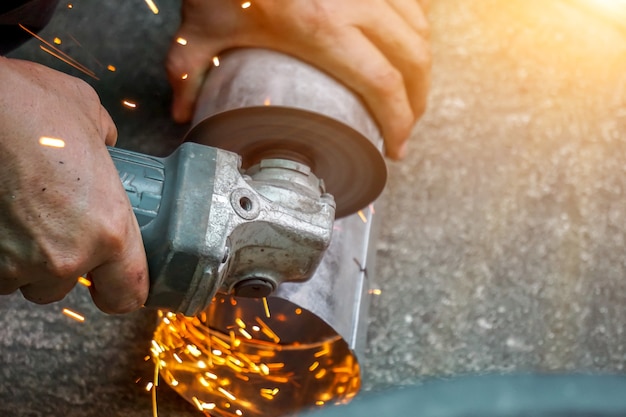 Photo closeup hands of laborer holding electric angle grinder working cutting galvanized pipe at construction site with sun flare background.