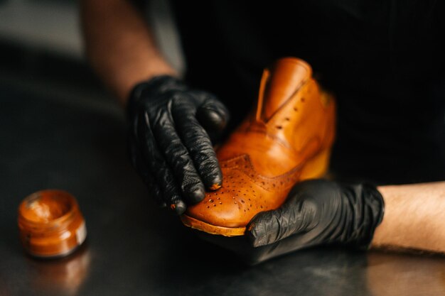 Closeup hands of shoemaker in black gloves rubbing paint on toe cup of light brown leather shoes