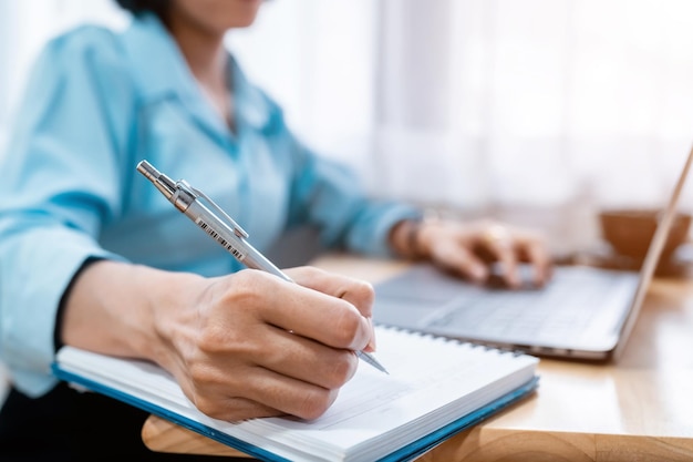 Closeup of hands Taking written Notes to on notebook and freelance business female casual working with laptop computer warm milk cup in coffee shop likeDistance job communication concept