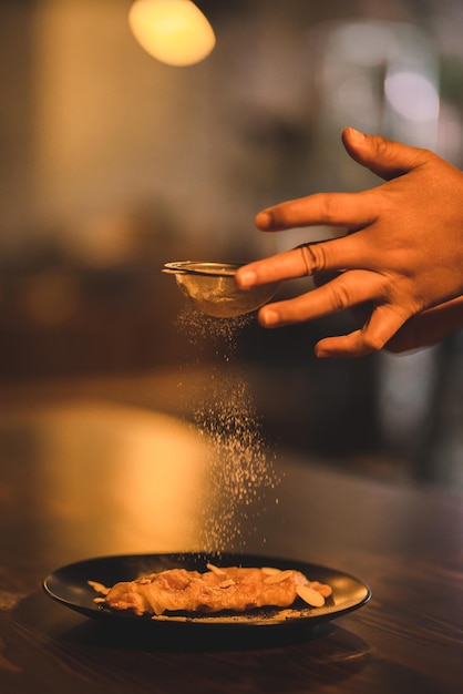 Closeup of hands of young chef sprinkling powdered sugar on sweet dish served in a black plate with nuts