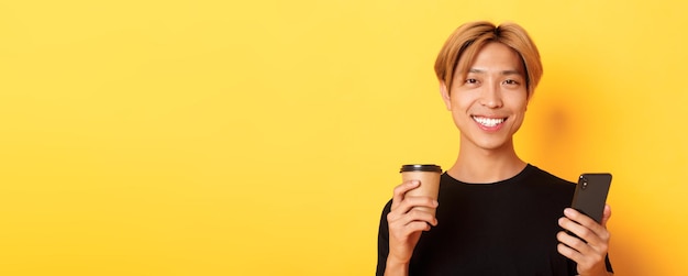 Closeup of handsome young asian guy smiling happy using smartphone and drinking takeaway coffee stan