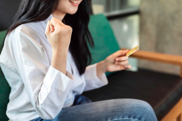 Крупным планом счастливая азиатка подняла кулаки и держит кредитную карту с успешными покупками в Интернете