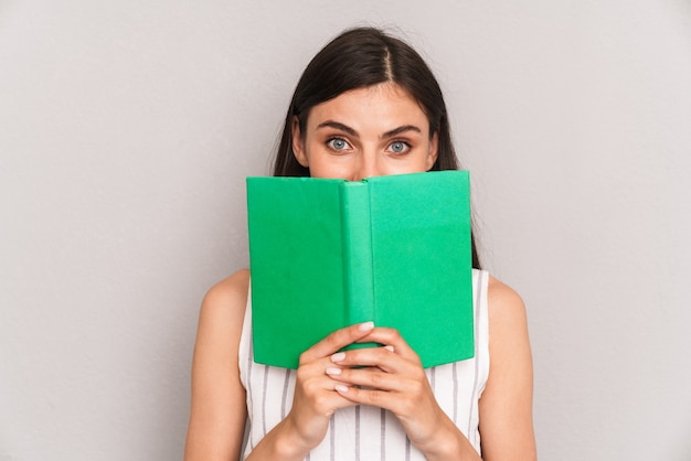 closeup of happy brunette woman wearing dress and covering her face with book isolated over gray wall