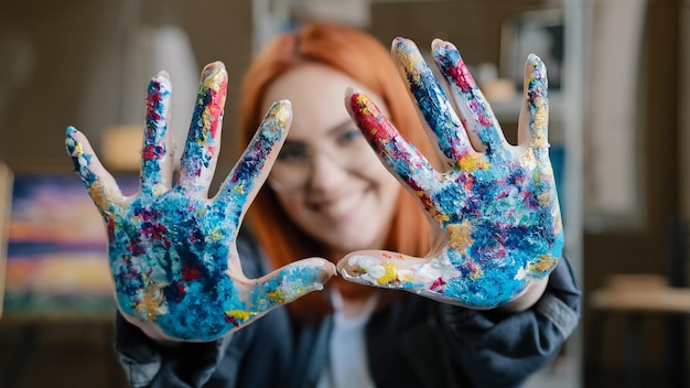 Photo closeup happy funy woman with red hair painter girl artist in glasses looks at camera stretches