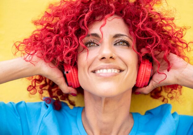 Closeup of a happy latin woman with red afro hair listening to music with her red headphones holding them with her hands