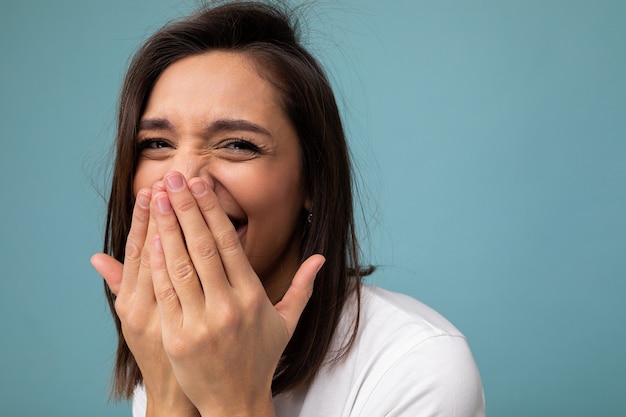 Closeup of happy positive young cute nice brunette woman with sincere emotions wearing casual white t-shirt for mockup isolated on blue background with copy space and laughing covering mouth.