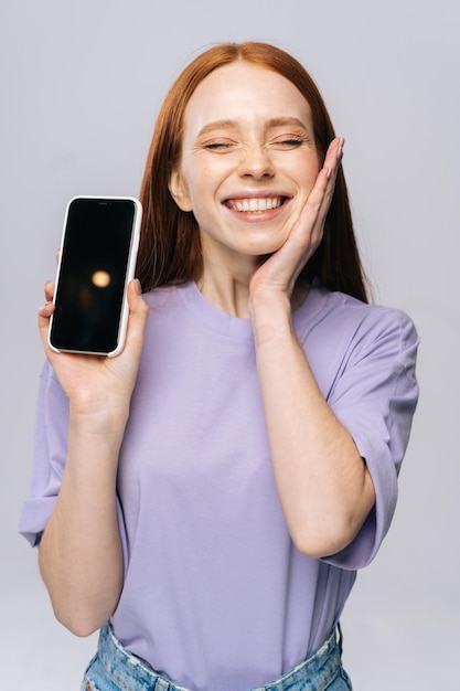 Closeup of happy young woman in casual clothes showing phone with black empty mobile screen holding hand near face Pretty lady model with red hair emotionally showing facial expressions in studio