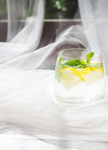 Closeup of homemade lemonade with lemon mint and ice cubes in a glass on white wood table with white tulle fabric