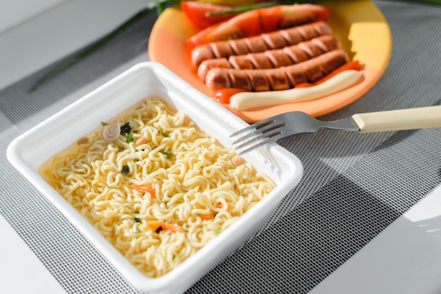 Closeup hot instant noodles fried sausages with ketchup and mayonnaise green onions and tomatoes on table