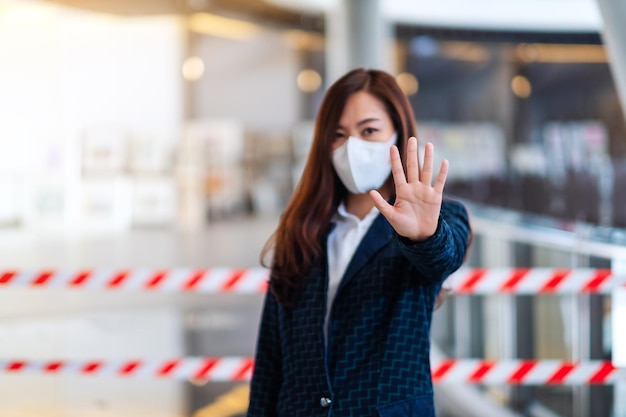 保護フェイスマスクを着用し、パンデミックの概念であるCovid-19の蔓延を防ぐために、赤と白の警告テープ領域の前にストップハンドサインを作成しているアジア人女性のクローズアップ画像