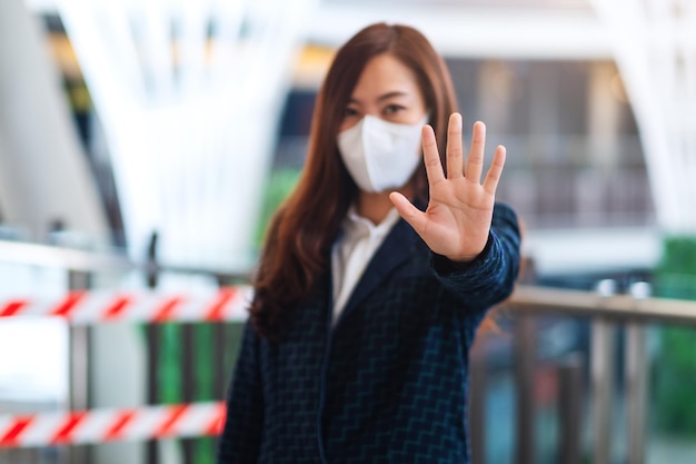 保護フェイスマスクを着用し、パンデミックの概念であるCovid-19の蔓延を防ぐために、赤と白の警告テープ領域の前にストップハンドサインを作成しているアジア人女性のクローズアップ画像