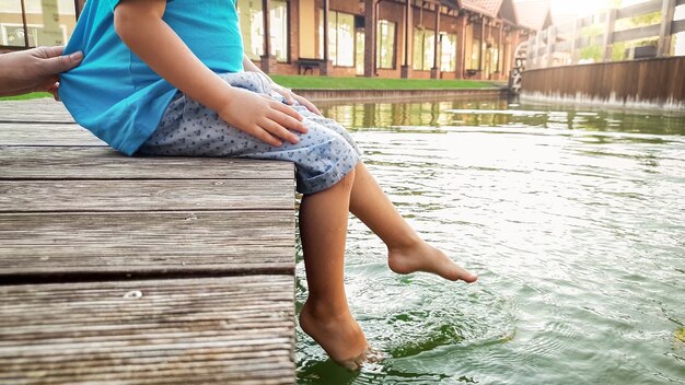 湖の木製の橋の上に座って、足で水をはねかける裸足の小さな幼児の少年のクローズアップ画像