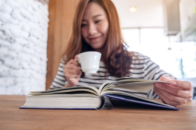 Closeup image of a beautiful asian woman holding and reading a book while drinking coffee