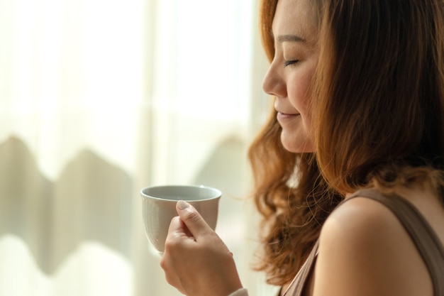 Immagine del primo piano di una bella donna asiatica che tiene e che sente l'odore di una tazza di caffè caldo con la sensazione rilassata al mattino