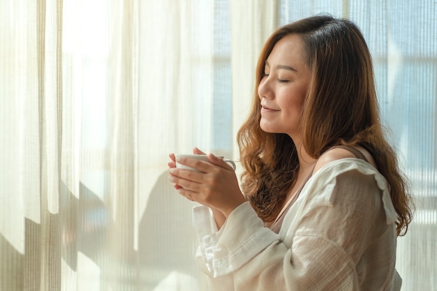 Immagine del primo piano di una bella donna asiatica che tiene e che sente l'odore di una tazza di caffè caldo con la sensazione rilassata al mattino