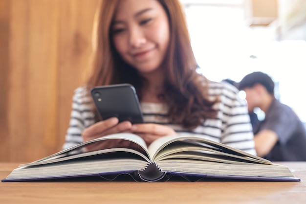 Closeup image of a beautiful asian woman holding , using and looking at smart phone with a book on wooden table