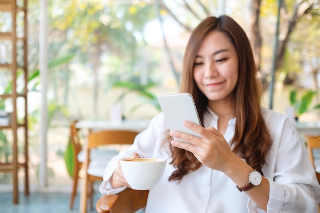 カフェでコーヒーを飲みながら携帯電話を持って使用している美しいアジアの女性のクローズアップ画像