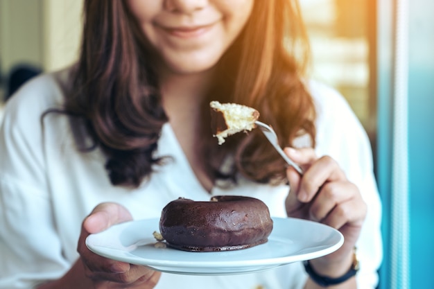 Крупным планом изображение красивой женщины, держащей и использующей вилку, чтобы съесть шоколадный пончик в белой тарелке