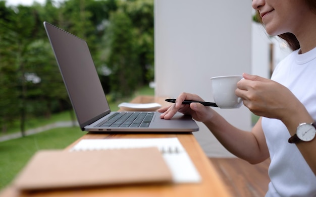 Closeup image of a beautiful young asian woman drinking coffee while using and working on laptop computer in the outdoors