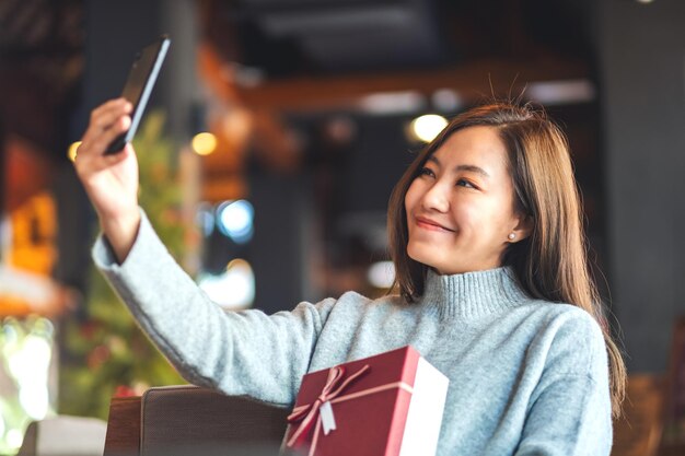 Closeup image of a beautiful young asian woman using mobile phone to take selfie with gift box