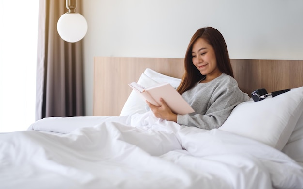 Closeup image of a beautiful young woman reading book in a white cozy bed at home