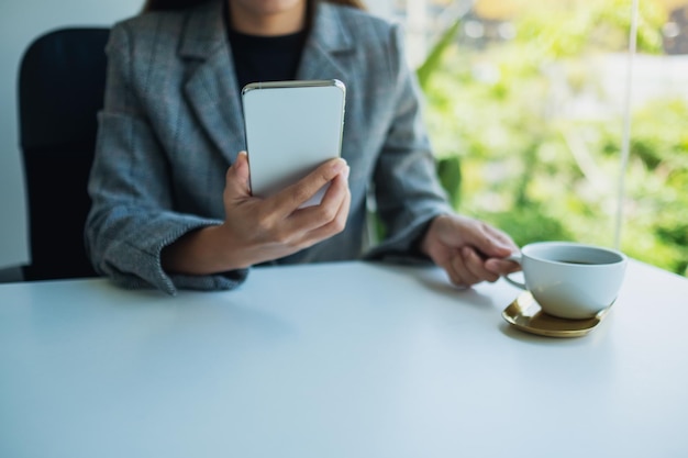 Closeup image of a businesswoman holding and using mobile phone while drinking coffee in office