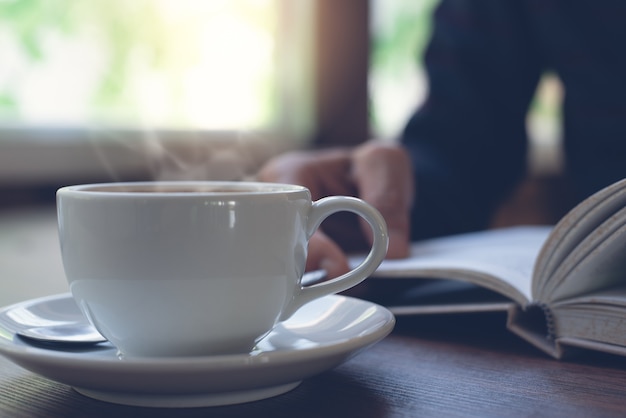 Closeup image of cup of coffee with man reading a book as background