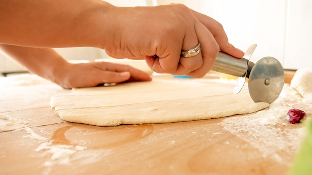 Immagine del primo piano della mano femminile che tiene il coltello rotondo per pizza e taglia la pasta su una grande scrivania di legno in cucina