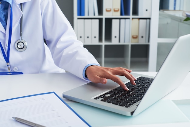 Крупным планом изображение врача общей практики, работающего на ноутбуке и подписывающего документы за ее офисным столом
