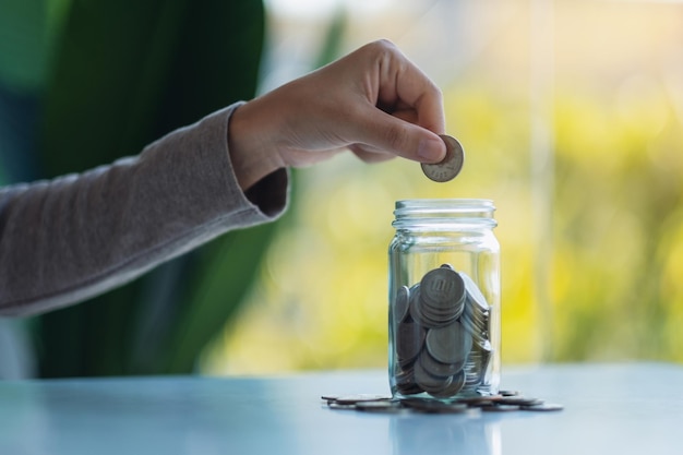 Closeup image of a hand collecting and putting coins in a glass jar for saving money concept