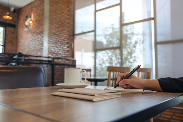 Closeup image of a hand writing on blank notebook with coffee cup on table in cafe