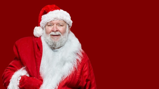 Изображение крупного плана счастливого смеющегося Санта-Клауса, одетого в красное пальто и шляпу, держит сумку на спине, изолированном красном фоне. Место для текста.