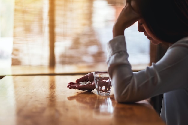 Photo closeup image of a headache woman holding a white pill with a glass of water on the table