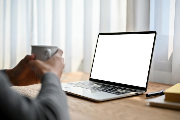 Closeup image of a man holding a cup of coffee while looking at laptop screen laptop mockup