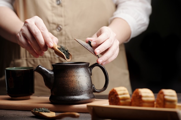 Closeup image of man putting spoon of dried tea leaves in ceramic pot