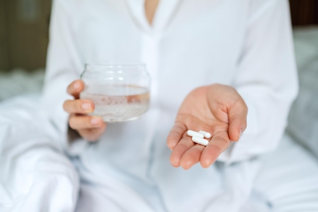 Closeup image of a sick woman holding white pills and a glass of water while sitting on a bed