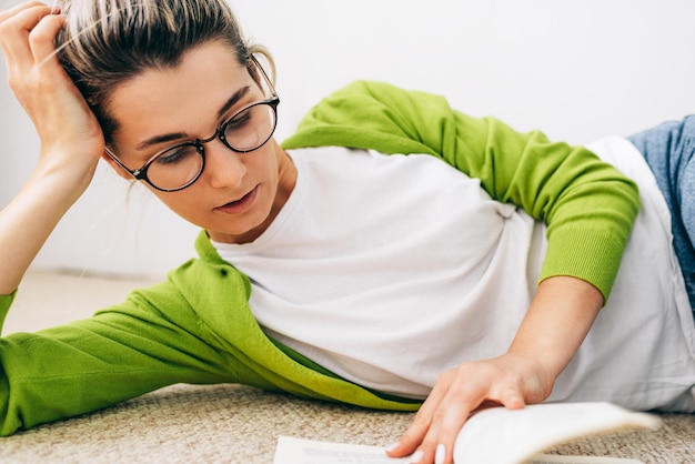 Closeup image of smart young woman reading book wearing green cardigan and white tshirt eyeglasses lying on the carpet Female relaxing during reading a book Student girl studying at home