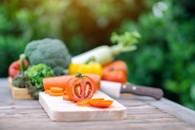 Closeup image of a tomato on chopping wooden board with knife and a tray of mixed vegetables on the table