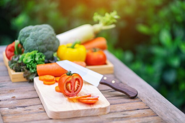 Closeup image of a tomato on chopping wooden board with knife and a tray of mixed vegetables on the table