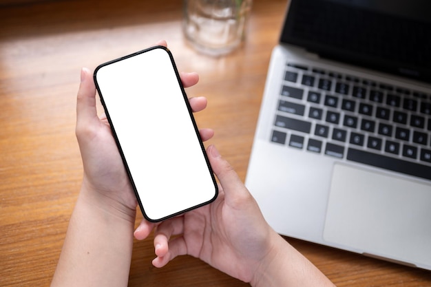 Closeup image of a whitescreen smartphone in a woman's hands above a wooden desk with a laptop