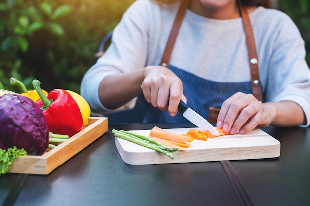 Closeup image of a woman cutting and chopping carrot by knife on wooden board with mixed vegetables in a tray