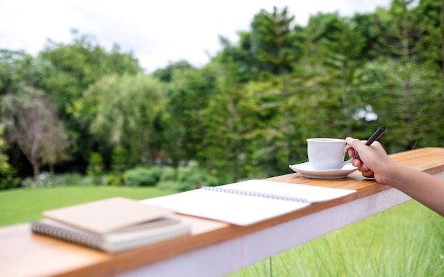 Closeup image of a woman drinking coffee and writing on a notebook in the outdoors